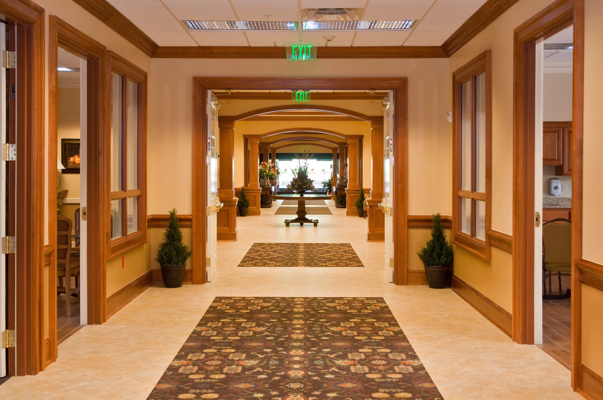 A long, well-lit hallway in a senior living facility with beige walls and wooden trim. The floor has a patterned carpet runner, and there are potted plants placed along the walls. Multiple doorways and windows line the hallway, and exit signs are visible on the ceiling. At the far end, there is a table with a floral arrangement.
