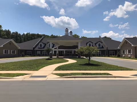Front exterior of a single-story senior living building with a covered drop-off porte-cochère, circular driveway, and lawn under a blue sky.