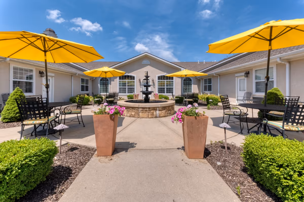 Outdoor courtyard area with a central stone fountain surrounded by patio tables and chairs with bright yellow umbrellas. The courtyard is bordered by a beige building with multiple windows and small green shrubs and flower pots with pink flowers.