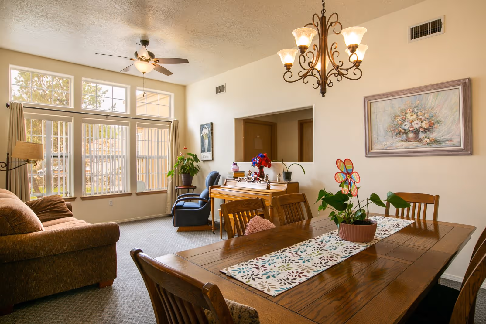 A cozy living and dining area with a wooden dining table and chairs, a brown sofa, a blue armchair, a piano with decorative items on top, large windows with vertical blinds, a ceiling fan, a chandelier, and framed artwork on the walls.