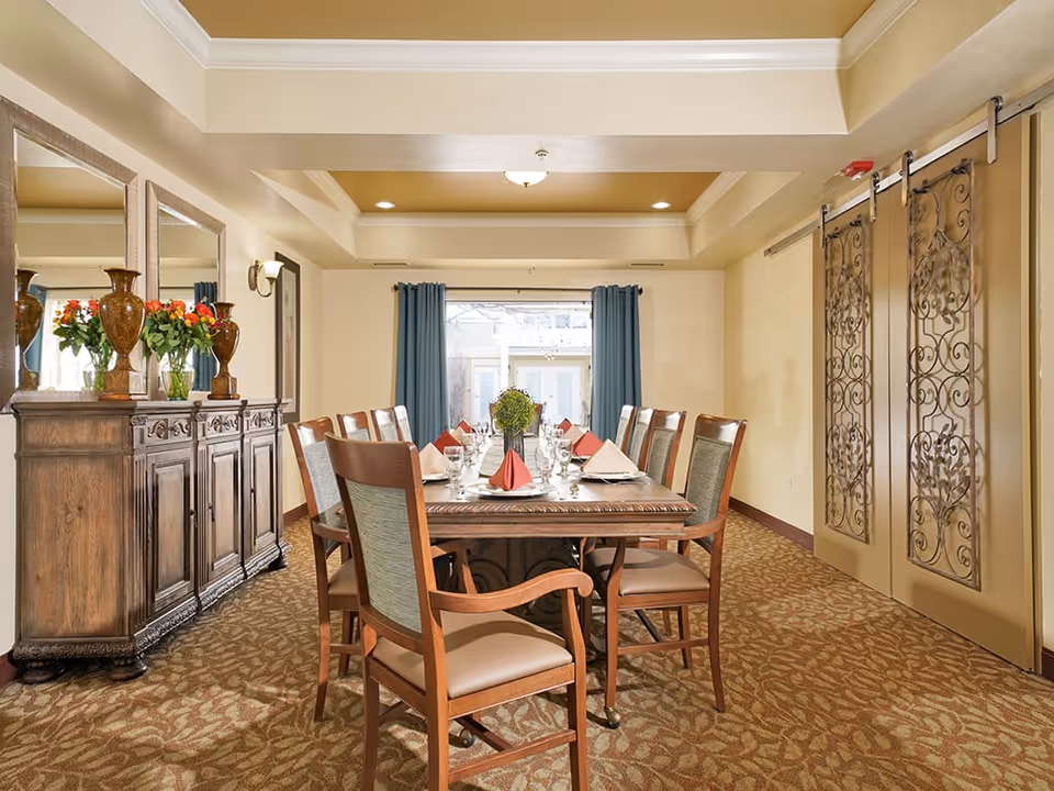A formal dining room with a long wooden table set for eight people. The table is decorated with folded red and white napkins, glassware, and a centerpiece of greenery. The room features patterned carpet, a wooden sideboard with vases and flowers, three large mirrors above the sideboard, blue curtains on a window at the far end, and decorative sliding doors with intricate metalwork on the right side.
