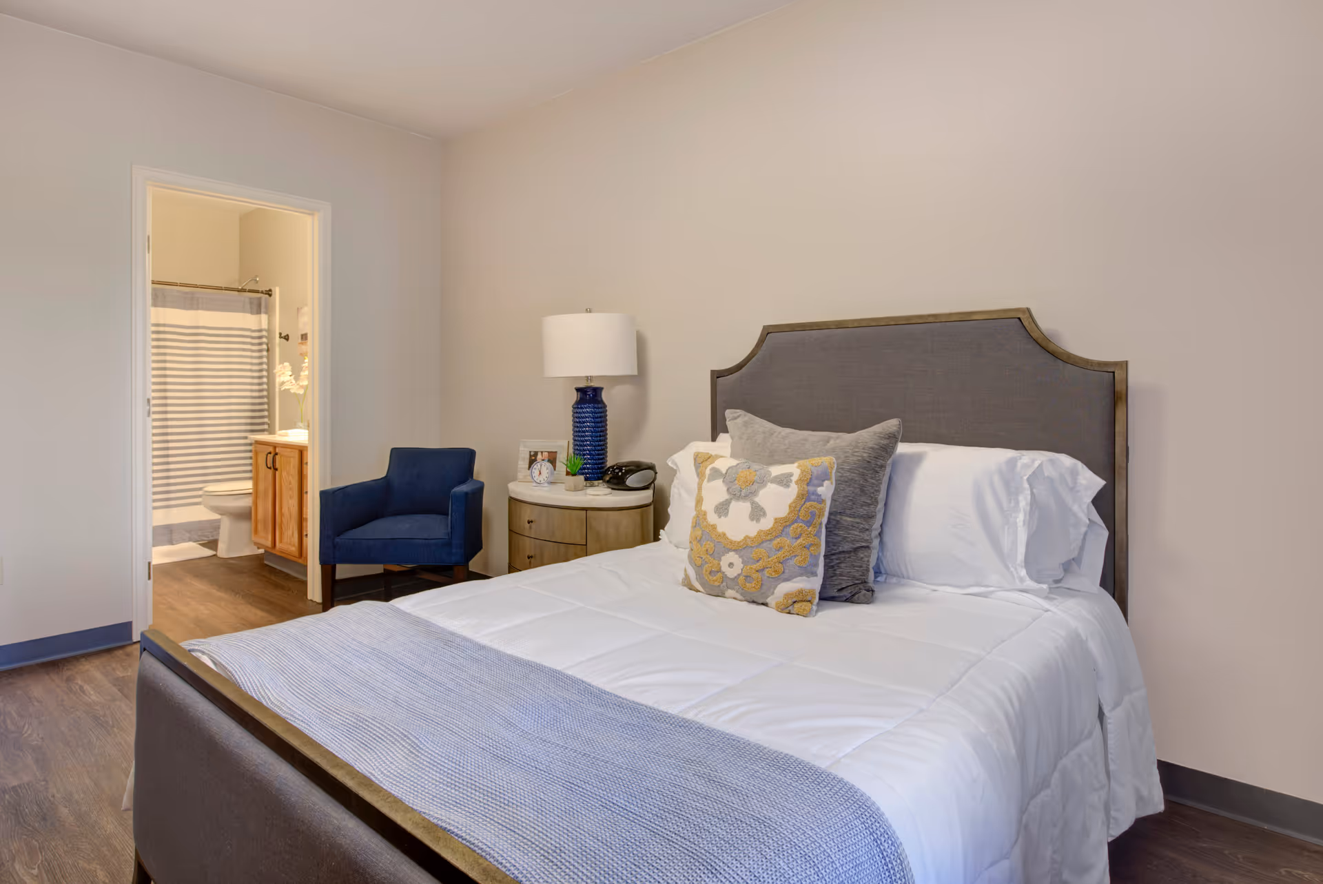 A furnished bedroom with a made bed, decorative pillows, a bedside table and lamp, a blue chair, and an open door showing an en-suite bathroom.