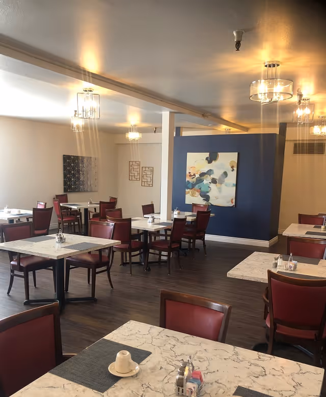 A dining room with multiple square marble-patterned tables and red cushioned chairs. The room has wooden flooring, modern ceiling lights, and abstract artwork on the walls. Each table has a small condiment holder and placemats.