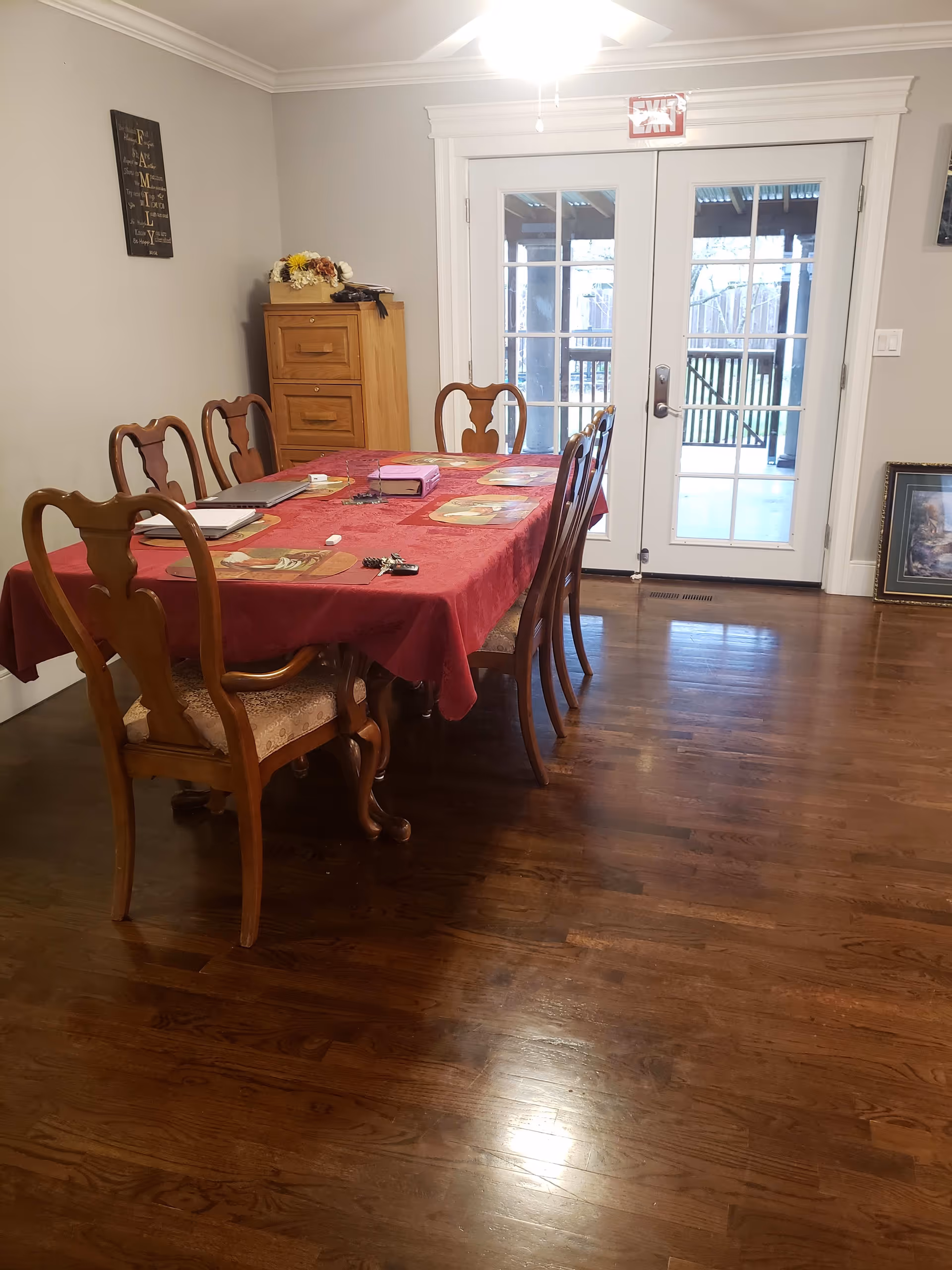A dining room with a wooden table covered by a red tablecloth and six wooden chairs. On the table are placemats, a laptop, a book, keys, and some small items. In the background, there is a wooden cabinet with decorative flowers on top and a wall decoration with the word FAMILY. Double glass doors lead to an outdoor area with a covered patio and a fenced yard.