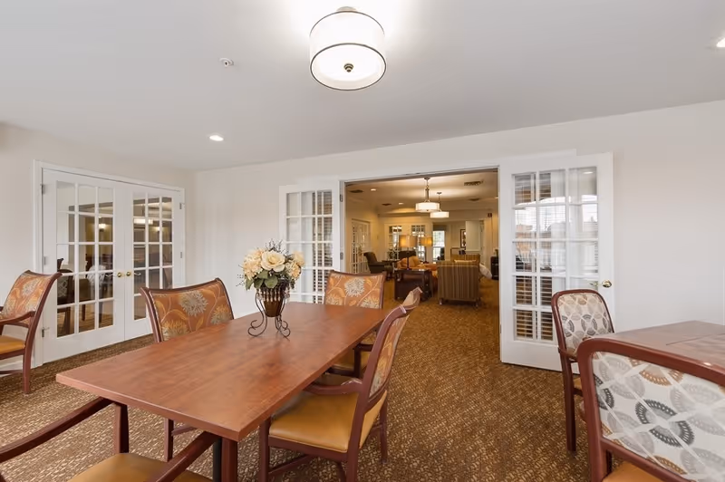 A dining area with a wooden table, floral centerpiece and upholstered chairs opening through glass French doors into a sitting room.