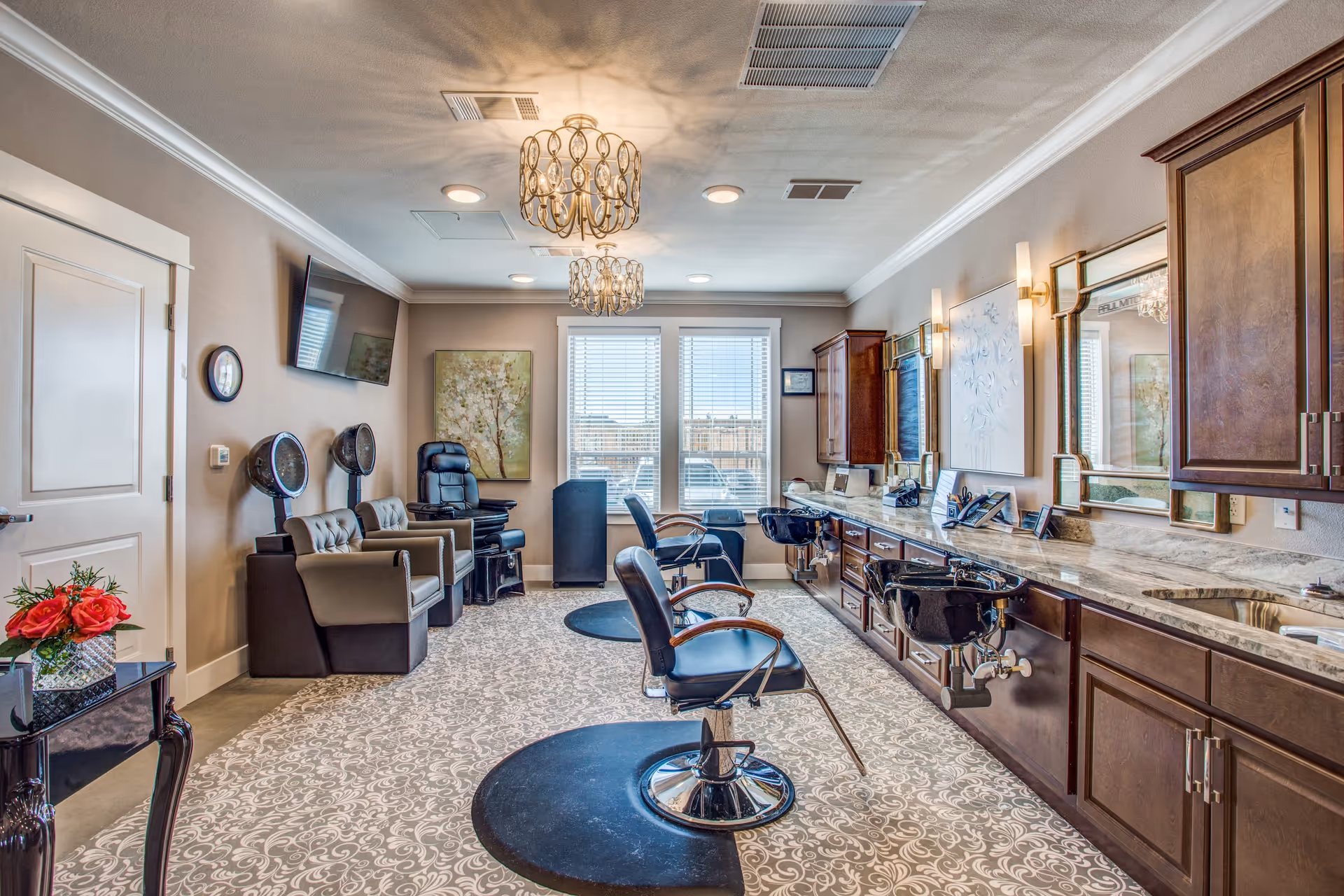 A well-lit salon room with two black salon chairs in front of sinks and mirrors on the right side. On the left side, there are two hair drying chairs with hooded dryers and a black massage chair near the window. The room has patterned carpet, beige walls, two decorative ceiling lights, and a small table with a vase of red flowers near the door.
