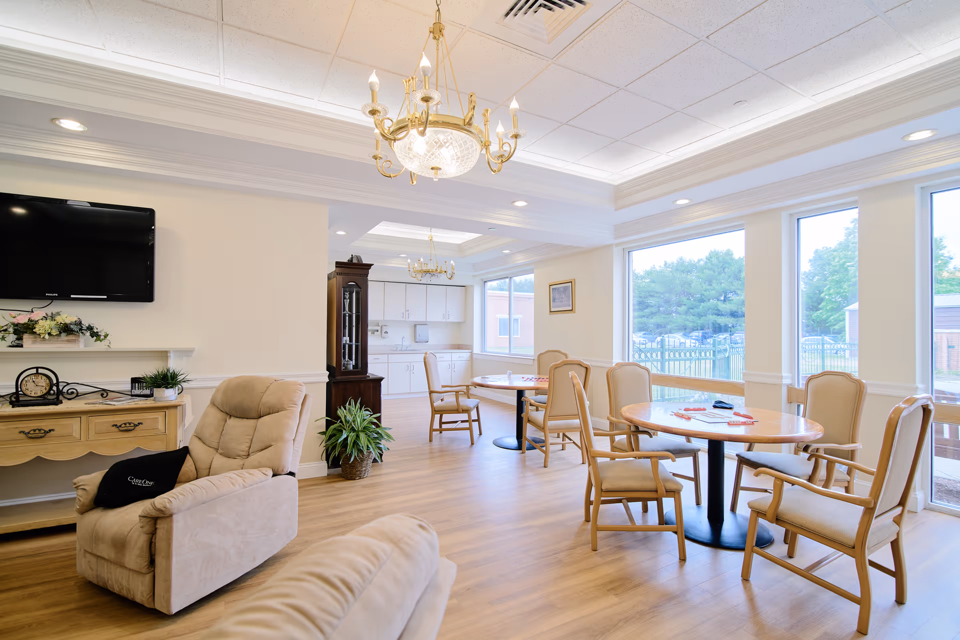 A bright and spacious common area in a senior living facility with beige cushioned chairs around wooden tables, large windows letting in natural light, a wall-mounted TV, a wooden cabinet, and a chandelier hanging from the ceiling. The room has light-colored walls and wood flooring.