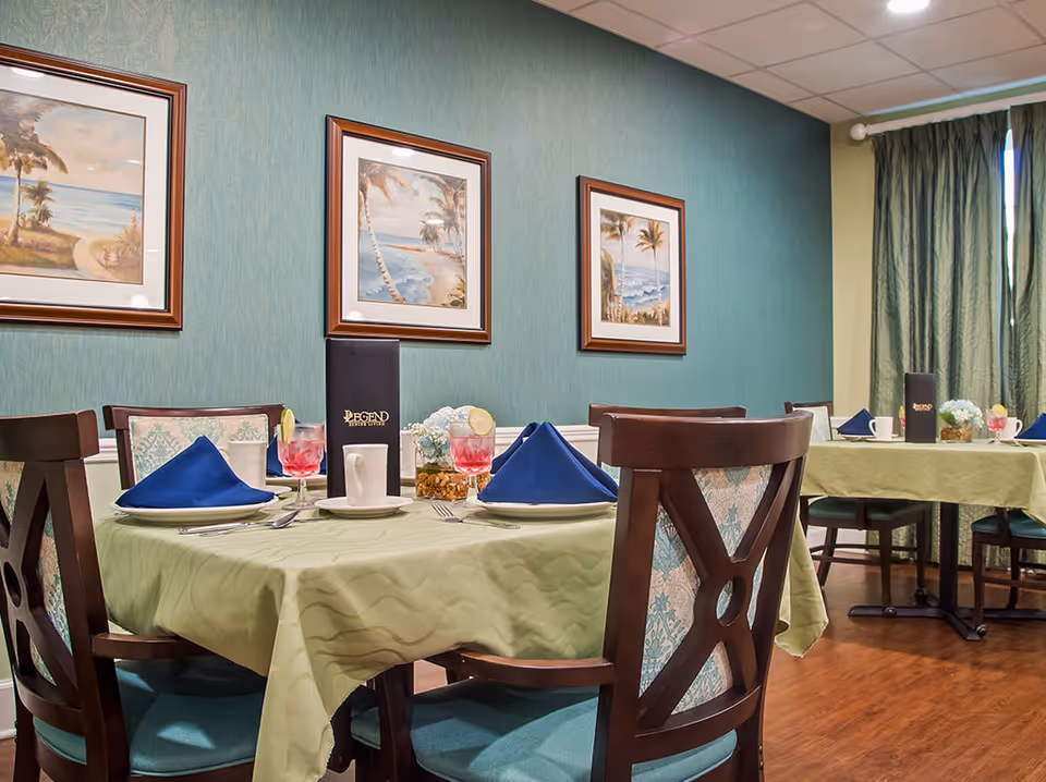Dining room with round tables draped in green tablecloths, place settings with blue napkins, wooden chairs, and framed beach paintings on teal walls.