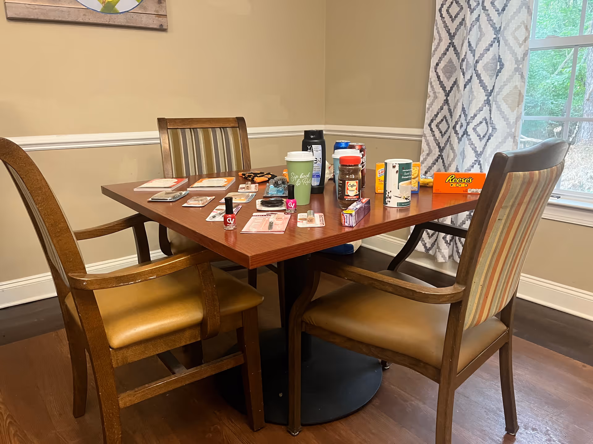 Wooden dining table with four chairs in a dining room, the table covered with snacks, drinks, and small personal items near a window.