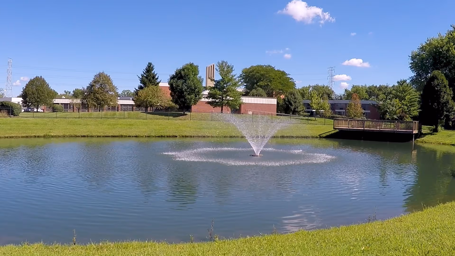 A pond with a central water fountain in front of a senior living facility building and grassy grounds under a blue sky.