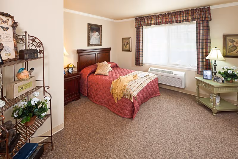 A cozy bedroom with a single bed covered in a red quilt and a beige throw blanket. The room has a wooden headboard, a nightstand with a lamp and clock, a window with plaid curtains, a green side table with a lamp and framed photos, and a metal shelf with decorative items and plants.