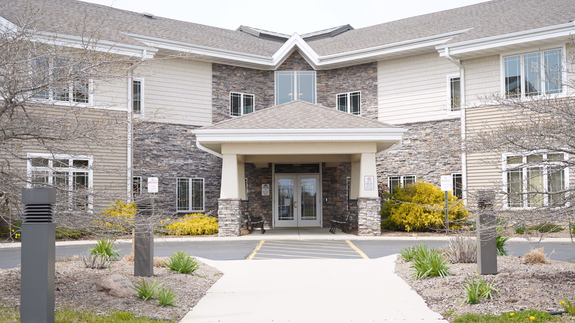 Front entrance of a two-story senior living building with a covered porte-cochere, stone facade, and landscaped walkway.