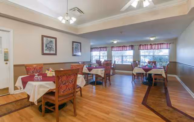 A dining room with several tables covered with white and burgundy tablecloths, each set with small flower arrangements. The room has wooden floors, large windows with valance curtains, framed artwork on the walls, and ceiling fans with lights.