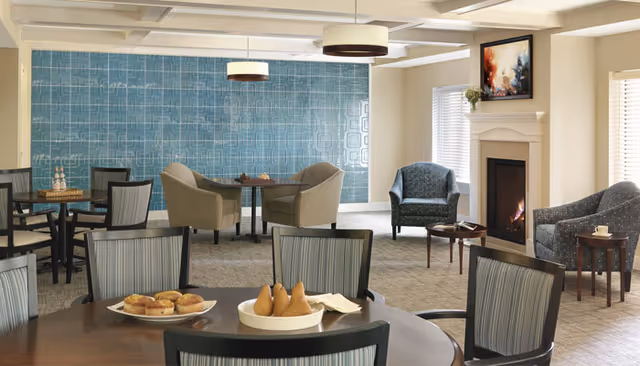 A cozy common area with several tables and chairs arranged for seating. The room features a fireplace with a painting above it, two armchairs near the fireplace, and a blue accent wall with a geometric pattern. On the tables, there are plates with muffins and pears. The space is well-lit with ceiling lights and natural light from windows with blinds.