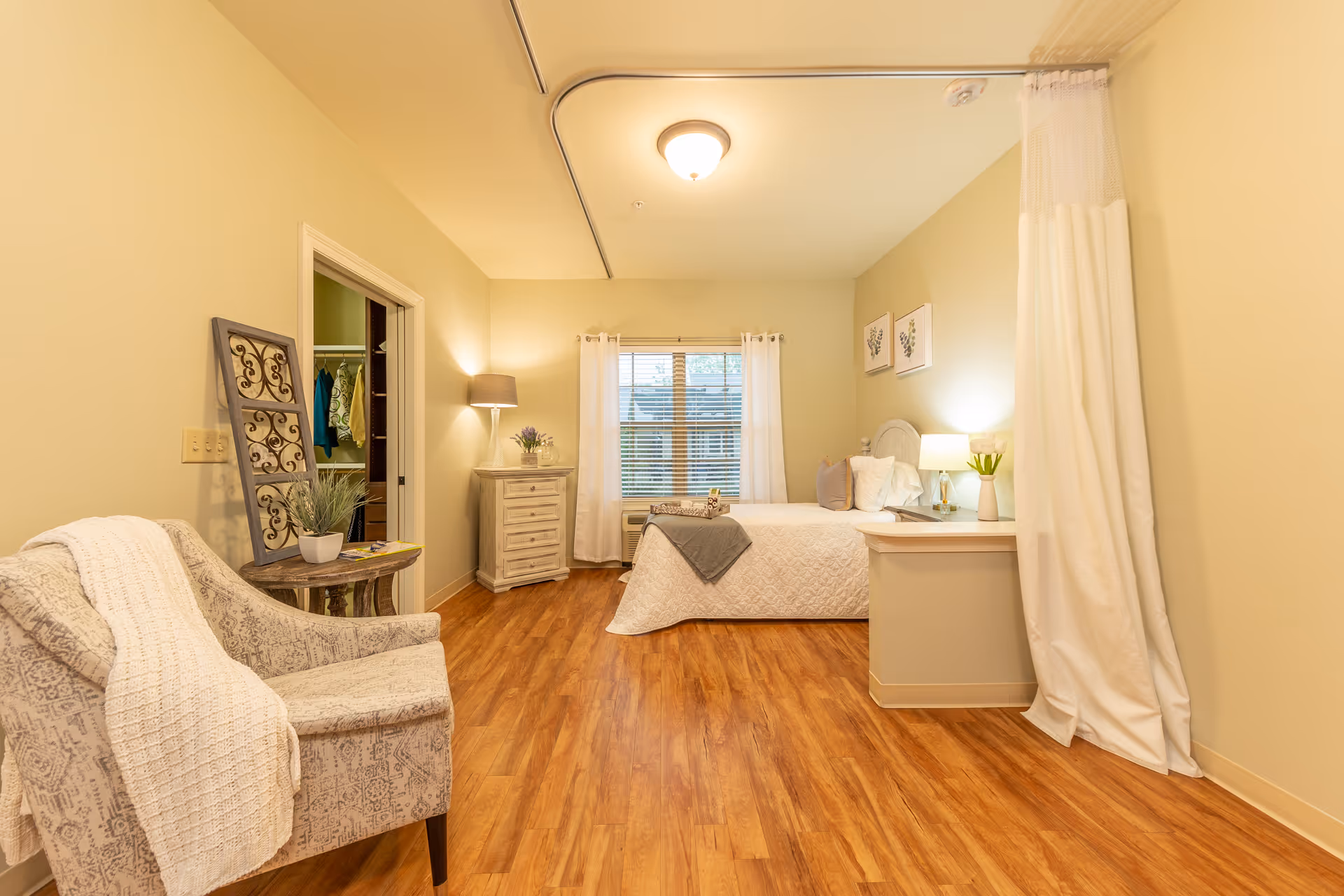 A cozy bedroom in Cedar Crest Memory Care featuring a bed with white bedding and pillows, a bedside table with a lamp and a plant, a window with white curtains, a small dresser with a lamp and flowers, a comfortable armchair with a white knitted throw, and a wooden floor. There is also a doorway leading to a closet with hanging clothes and a white privacy curtain on the right side.