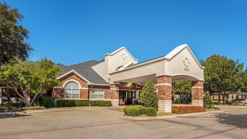 Exterior view of a senior living facility building with a covered entrance supported by brick columns, surrounded by trees and landscaping under a clear blue sky.
