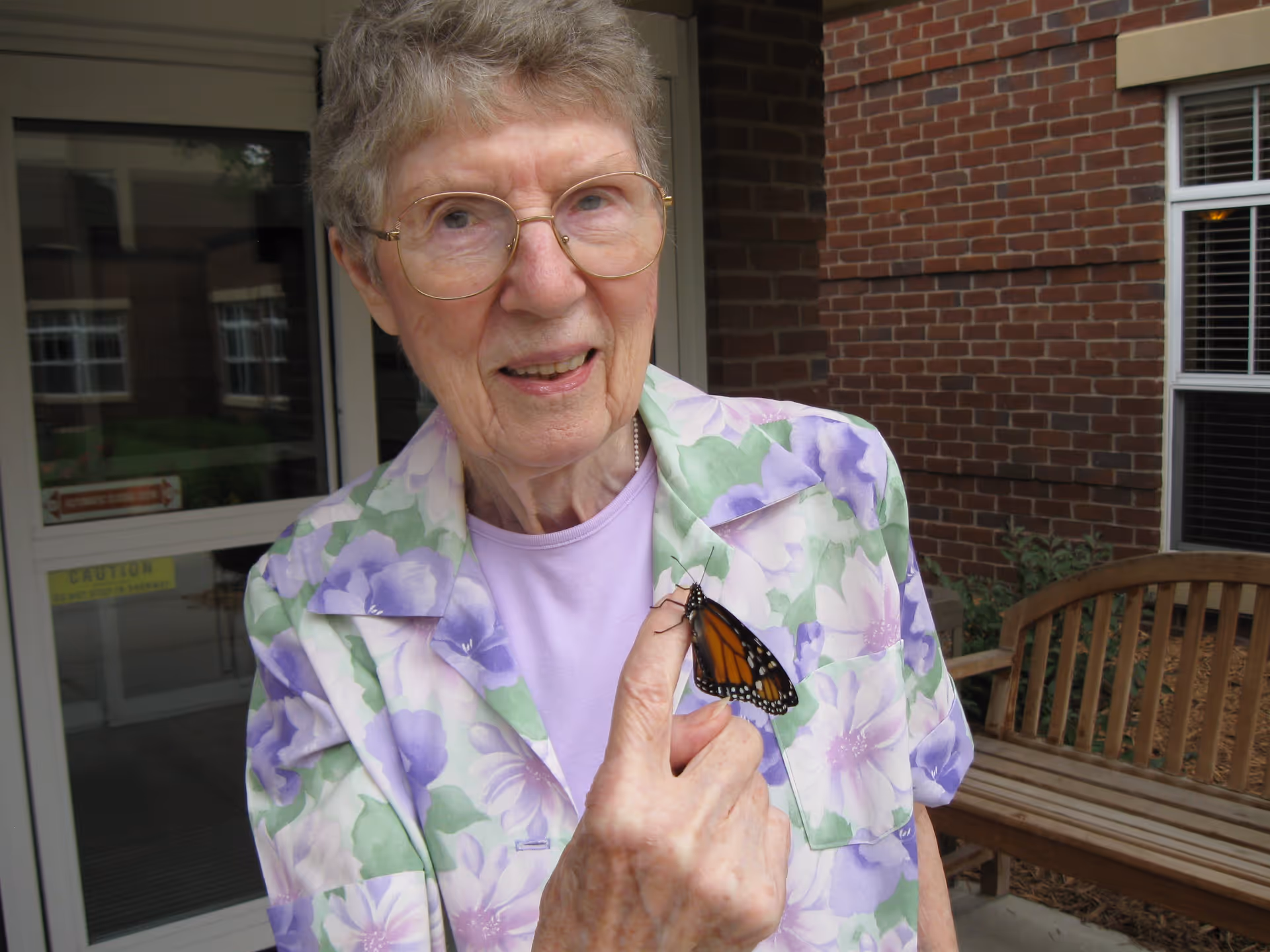 An elderly woman wearing glasses and a floral patterned jacket holds a monarch butterfly on her finger outside a brick building near a wooden bench.