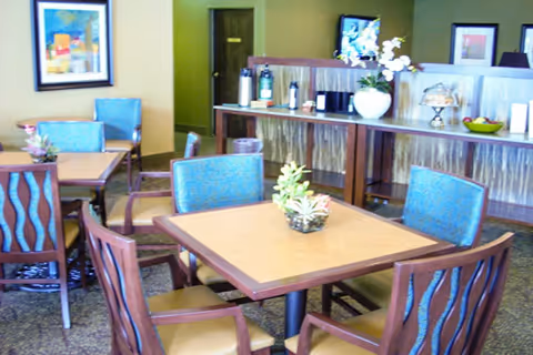 A dining area with square wooden tables and blue cushioned chairs. There are decorative plants on the tables and framed artwork on the walls. In the background, there is a counter with a white vase holding flowers and some other items, and a door leading to another room.