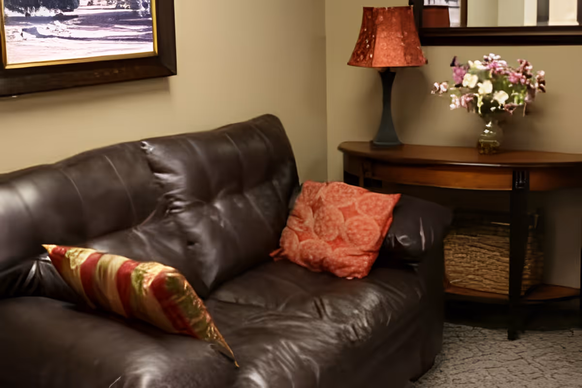A cozy living room area featuring a dark brown leather couch with two decorative pillows, one striped and one patterned. Behind the couch is a wooden side table with a red lampshade and a vase of flowers. A framed picture hangs on the wall above the couch.