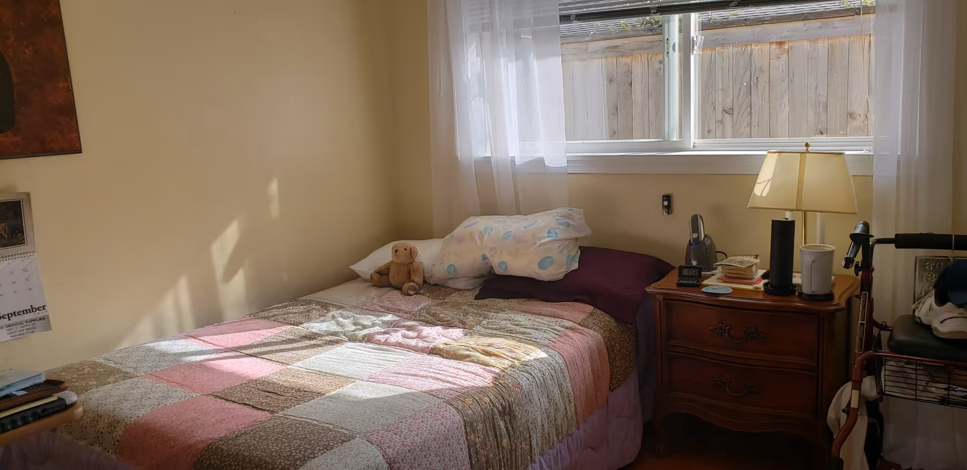 A small bedroom with a single bed covered in a patchwork quilt. There is a teddy bear on the bed along with pillows. Next to the bed is a wooden nightstand with a lamp, a cordless phone, some books, and a cup. A window with sheer white curtains lets in natural light. A walker is partially visible beside the nightstand.