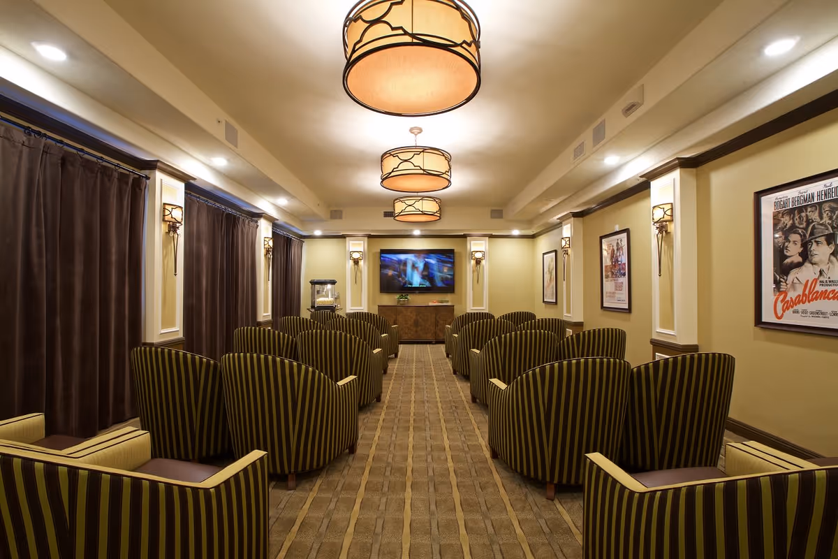 A cozy theater room with multiple rows of green and brown striped armchairs facing a wall-mounted flat screen TV. The room has warm lighting from ceiling fixtures and wall sconces, with dark curtains covering the windows and framed movie posters on the walls.