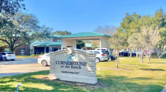 Exterior view of Cornerstone at the Ranch Bethany Rehabilitation facility showing a stone sign with the facility name and slogan 'Quality Loving Care' in front of a parking area with several cars and trees.