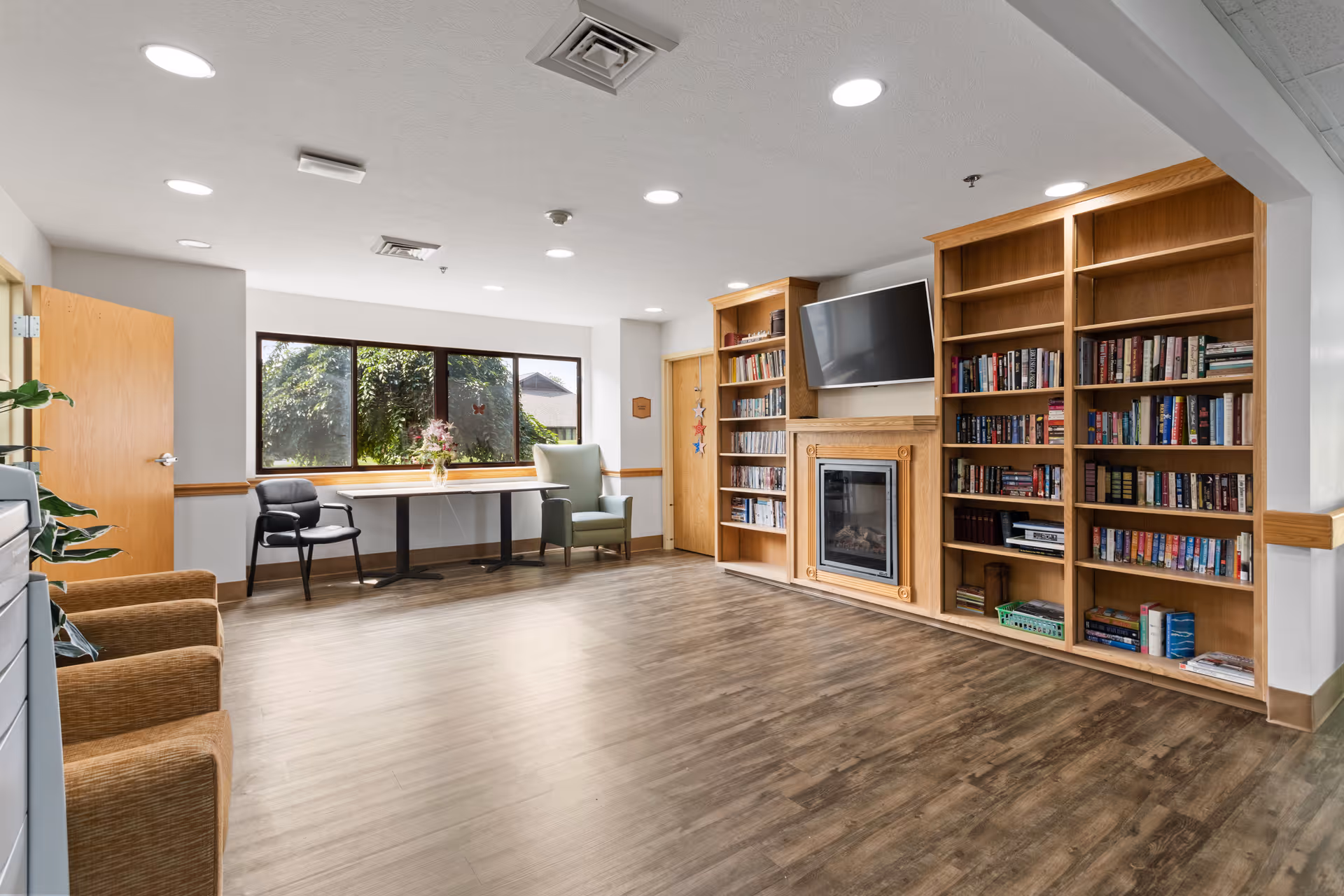 A bright and spacious room with wooden flooring, featuring built-in wooden bookshelves filled with books, a wall-mounted flat-screen TV above a decorative fireplace, a large window with a view of greenery outside, a table with two chairs near the window, and additional seating including armchairs and a black chair.