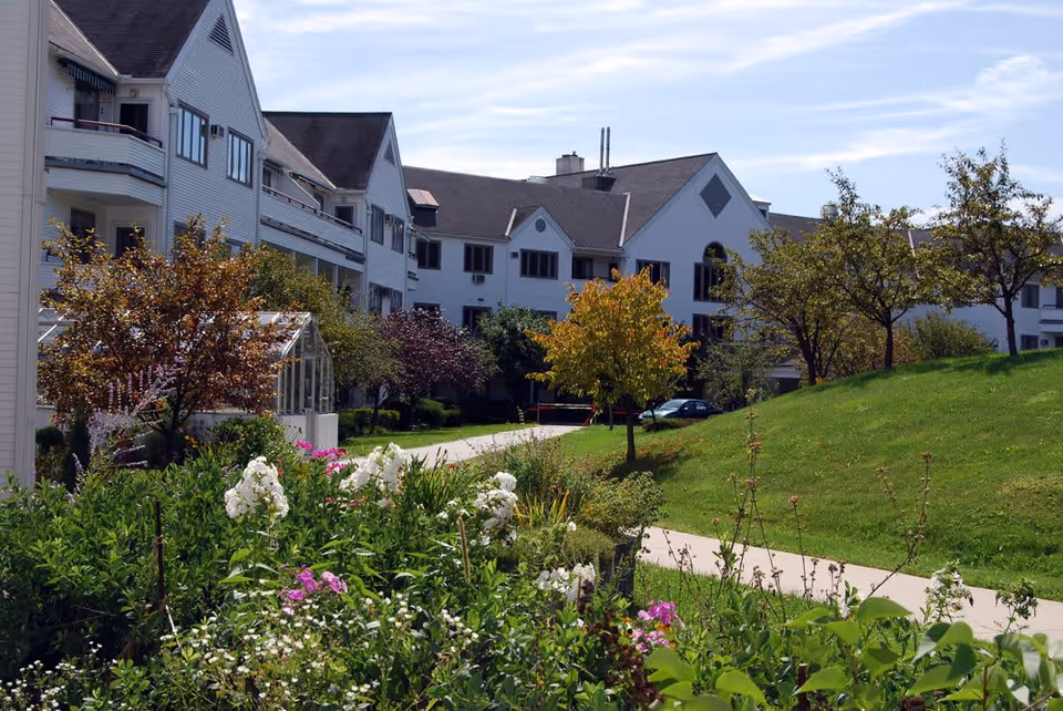 Exterior view of a senior living facility with white buildings, multiple windows, and balconies. The foreground features a garden with various flowering plants and shrubs, a paved walkway, and several trees with green and autumn-colored leaves under a partly cloudy sky.