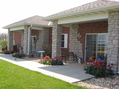 Senior living facility exterior with brick walls, stone columns, and a porch with blooming flowers.