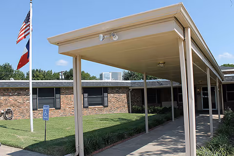 Covered walkway leading to the entrance of a single-story brick building with windows, a green lawn, and two flagpoles displaying the American and Texas flags under a clear blue sky.