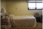 A small bedroom with a single bed covered by a beige crocheted blanket. There is a wooden nightstand with a lamp and some items on it next to the bed. A window with floral curtains is above a small chest of drawers on the right side of the room. The walls are painted light yellow.
