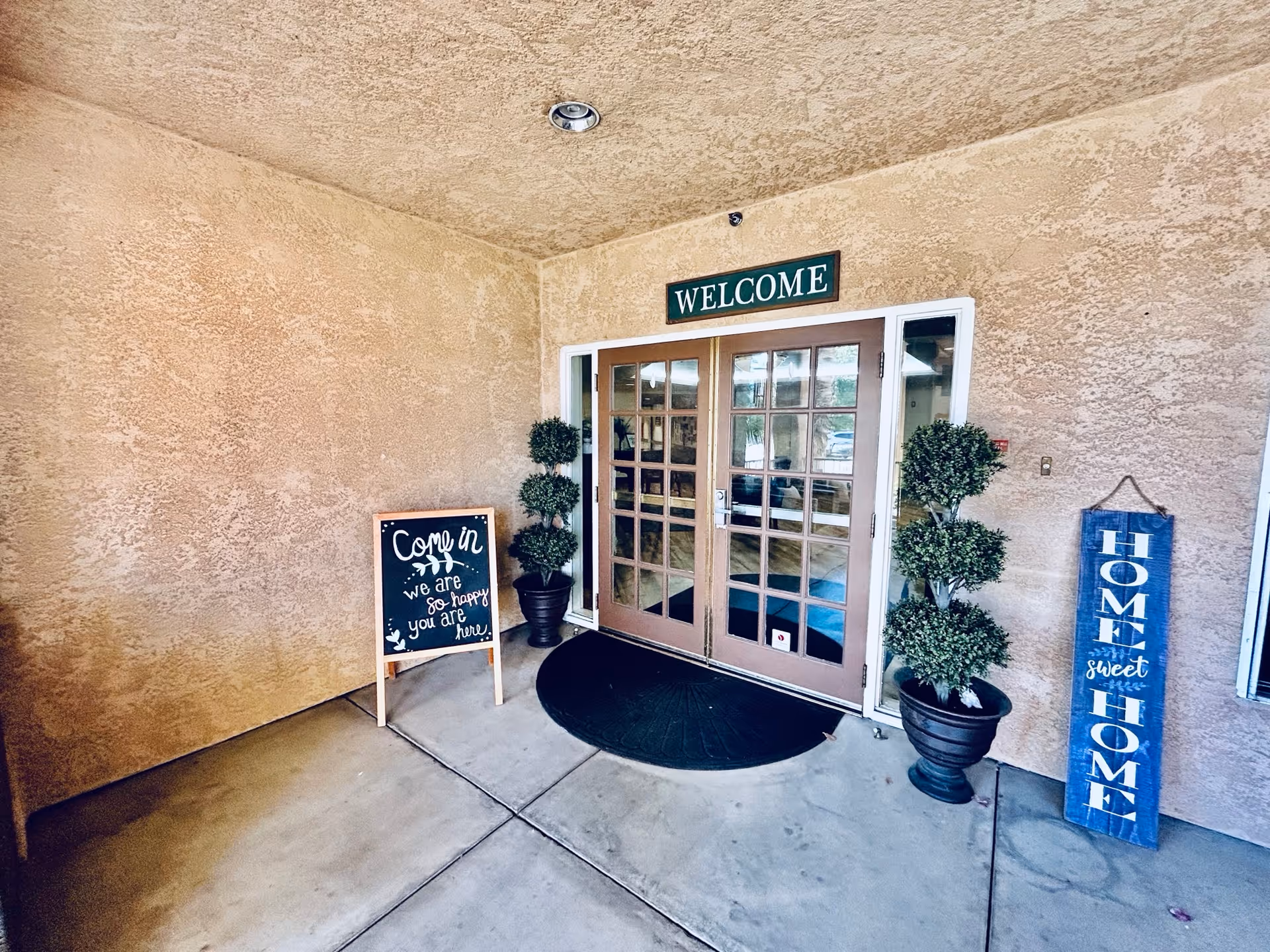 Entrance area of a facility with double glass doors under a 'WELCOME' sign. There are two potted topiary plants on either side of the doors. To the left, a chalkboard sign reads 'Come in we are so happy you are here.' To the right, a vertical wooden sign says 'HOME sweet HOME.' The walls and ceiling have a textured beige finish.