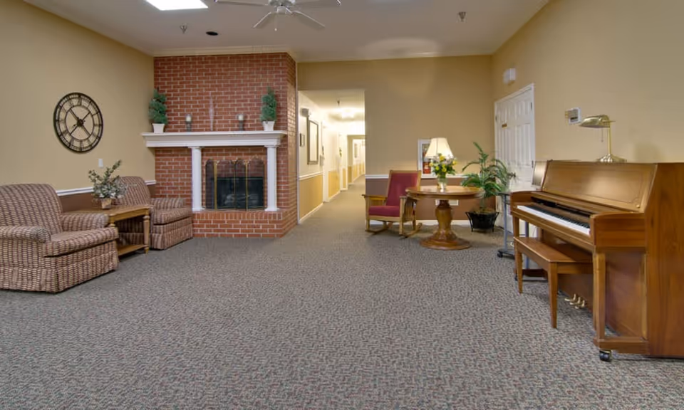 A cozy common area in a senior living facility featuring a brick fireplace with white mantel, two patterned armchairs with a small wooden table between them, a round wooden table with a lamp and flowers, a red armchair, a wooden piano with a bench, and a hallway leading to other rooms. The walls are painted beige with white trim, and the floor is carpeted with a patterned design.