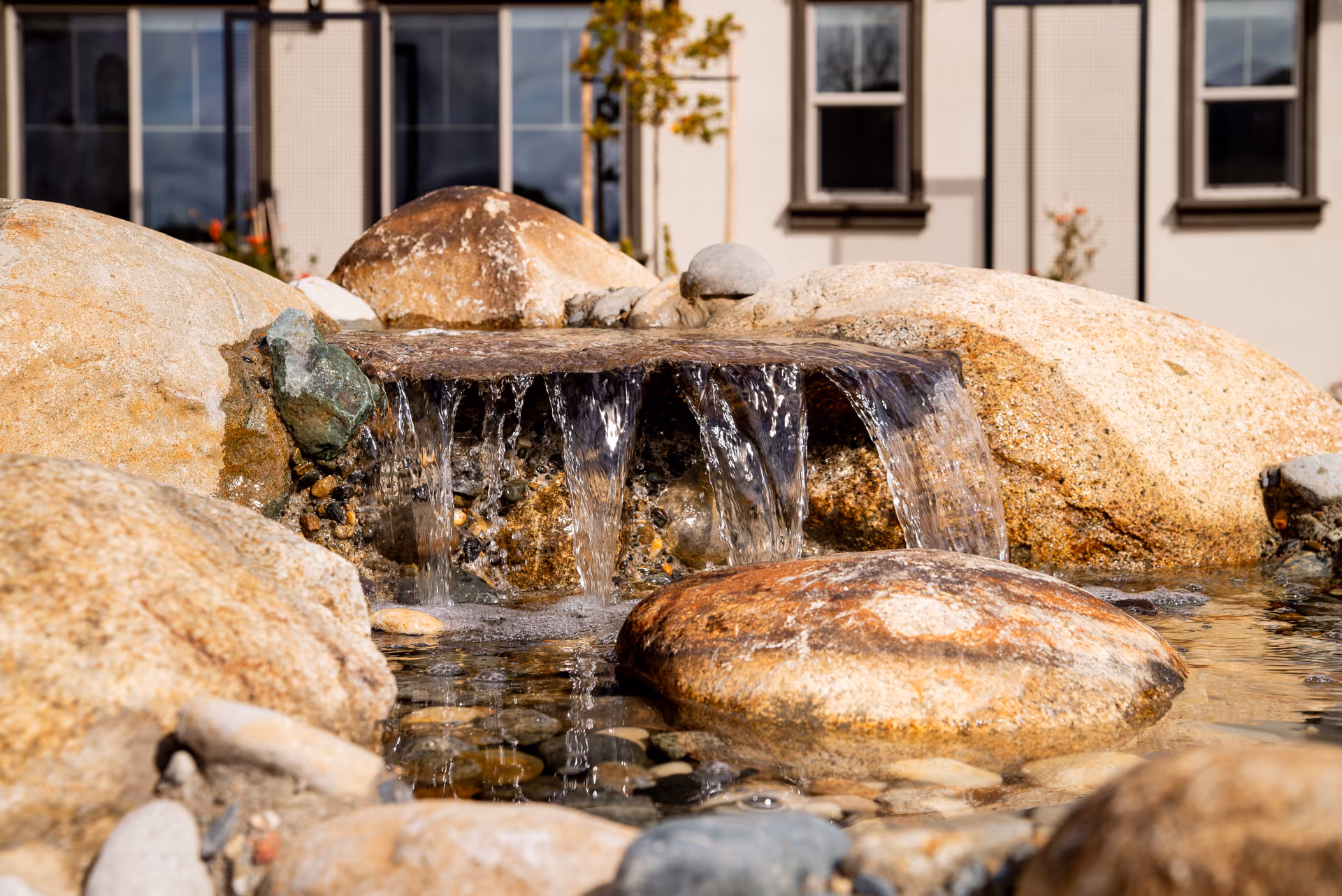 A close-up view of a small outdoor water feature with water cascading over flat rocks into a shallow pool surrounded by larger stones, with a building facade and windows visible in the background.
