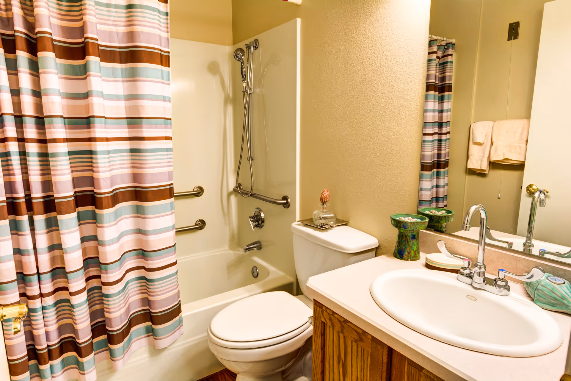 A bathroom with a bathtub and shower combination featuring a striped shower curtain in shades of brown, pink, and teal. There are two grab bars inside the bathtub area. A white toilet is positioned next to the bathtub. A countertop with a sink, faucet, and decorative items is visible, along with a mirror reflecting part of the shower curtain and a towel rack with beige towels.
