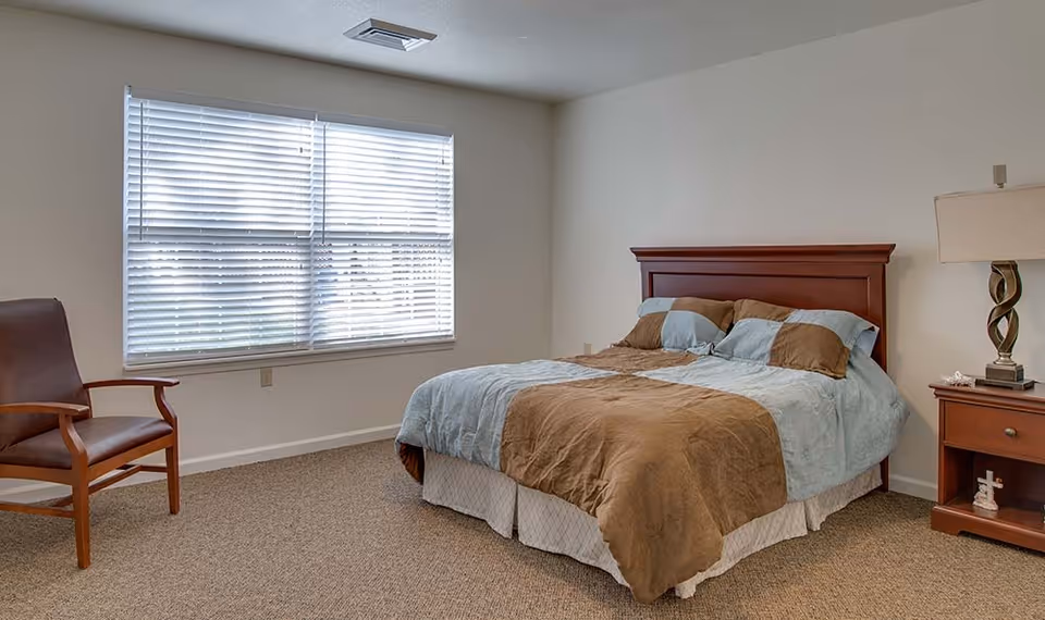 A bedroom with a large window covered by white blinds, a wooden bed with a brown and light blue comforter, a wooden nightstand with a lamp, and a wooden chair with armrests.