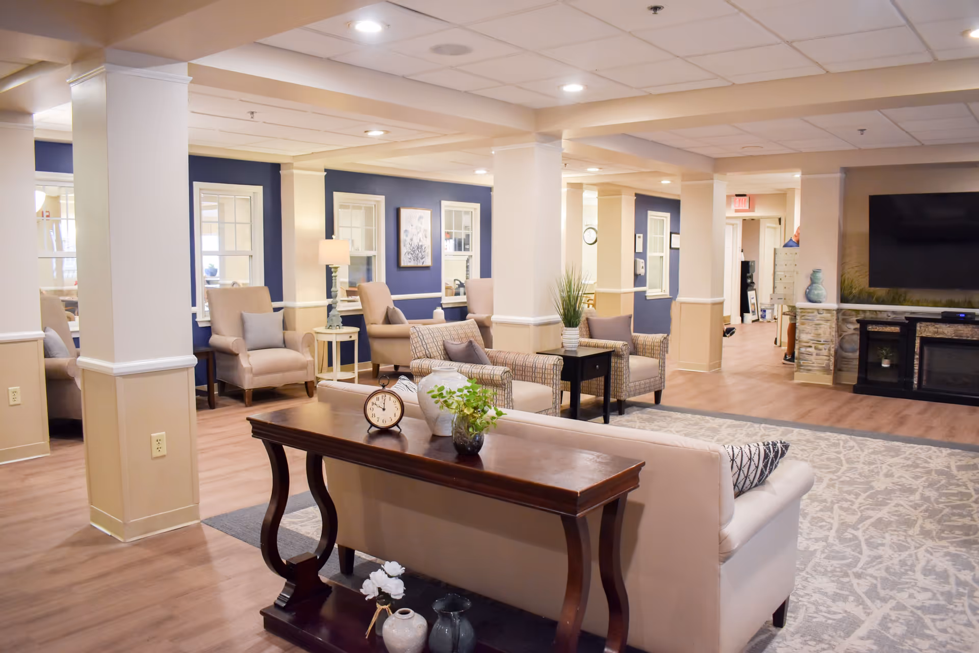 Well-lit communal living room with sofas, armchairs, side tables, a large TV over a fireplace, and decorative plants.