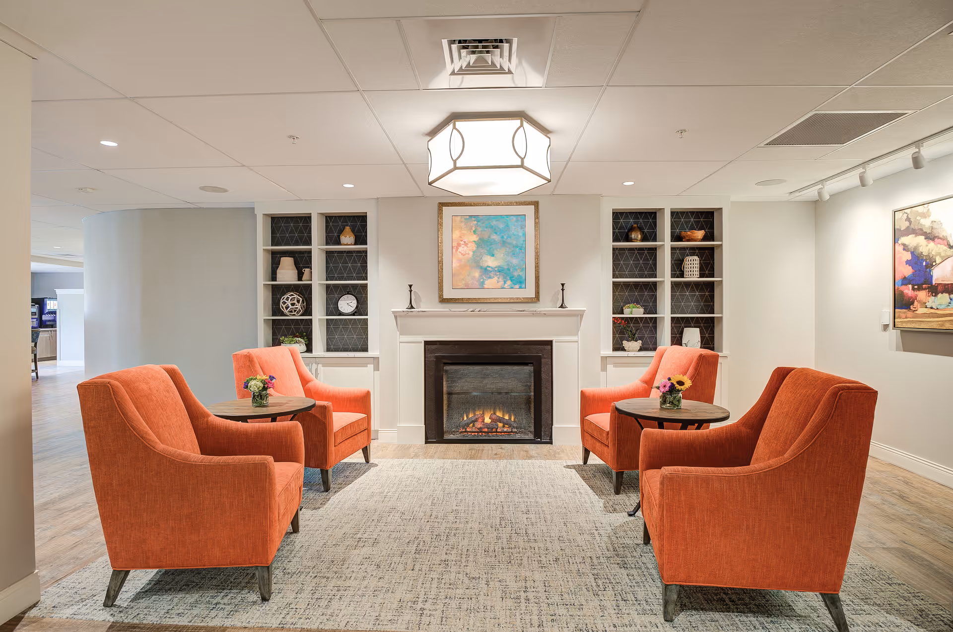 A cozy sitting area with four orange armchairs arranged around two small round tables, each with a small flower arrangement. The room features a fireplace with a colorful abstract painting above it, built-in shelves with decorative items on either side, and a modern ceiling light fixture. The walls are light-colored, and the floor has a light wood finish with a large area rug.