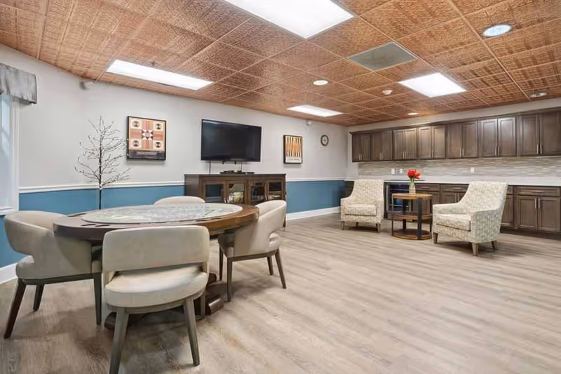 A spacious senior living common area with a round table surrounded by six cushioned chairs on the left side. On the right side, there are two patterned armchairs with a small round table between them holding a vase with red flowers. The back wall features dark wooden cabinets with a countertop and a mounted flat-screen TV above a wooden console. The room has a light wood floor, a blue and white painted wall, and a decorative wooden ceiling with recessed lighting.