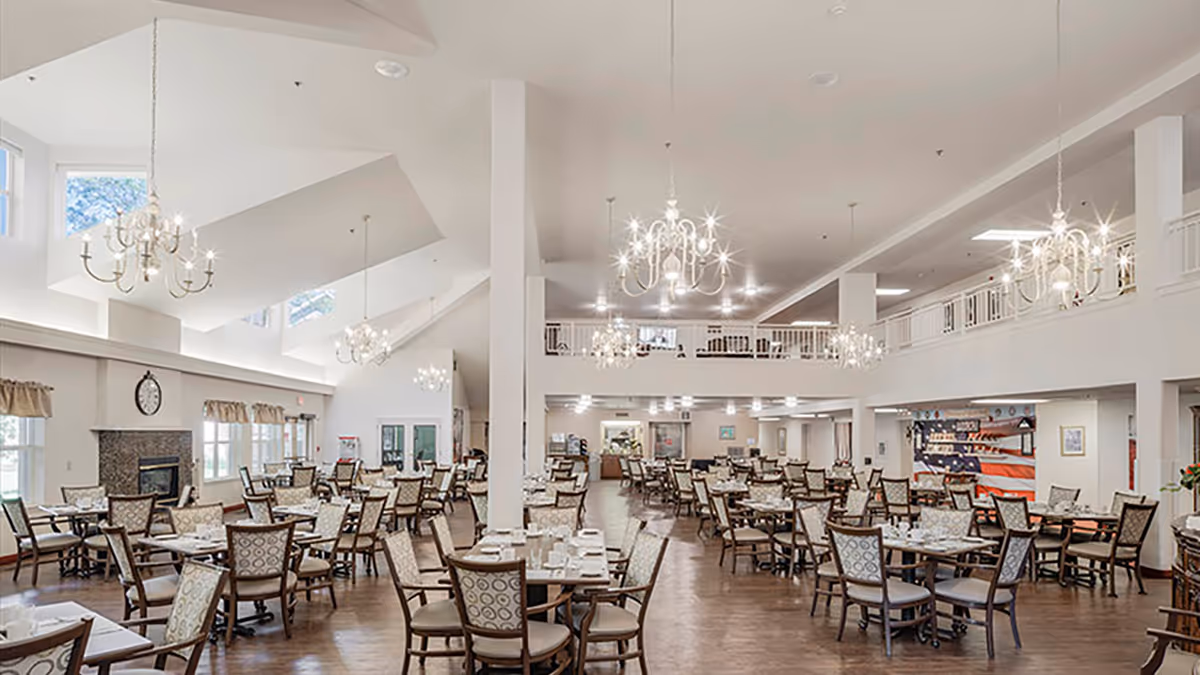 Spacious, well-lit dining room with many set tables, chandeliers, and a fireplace in a senior living facility.
