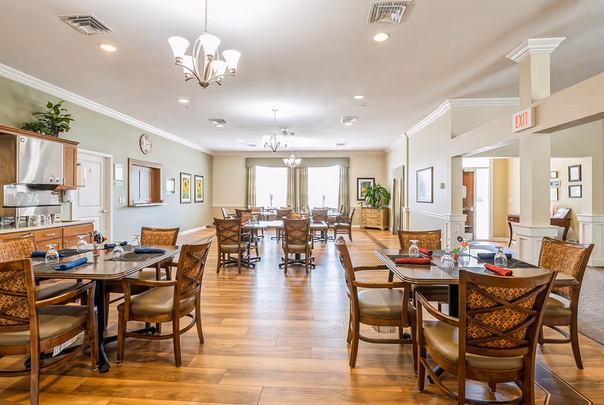 A bright and spacious dining room in an assisted living facility with wooden floors, multiple tables set with glasses and napkins, cushioned chairs, green walls with framed artwork, and ceiling light fixtures. There is a serving area with cabinets and a clock on the wall, and large windows with curtains letting in natural light.