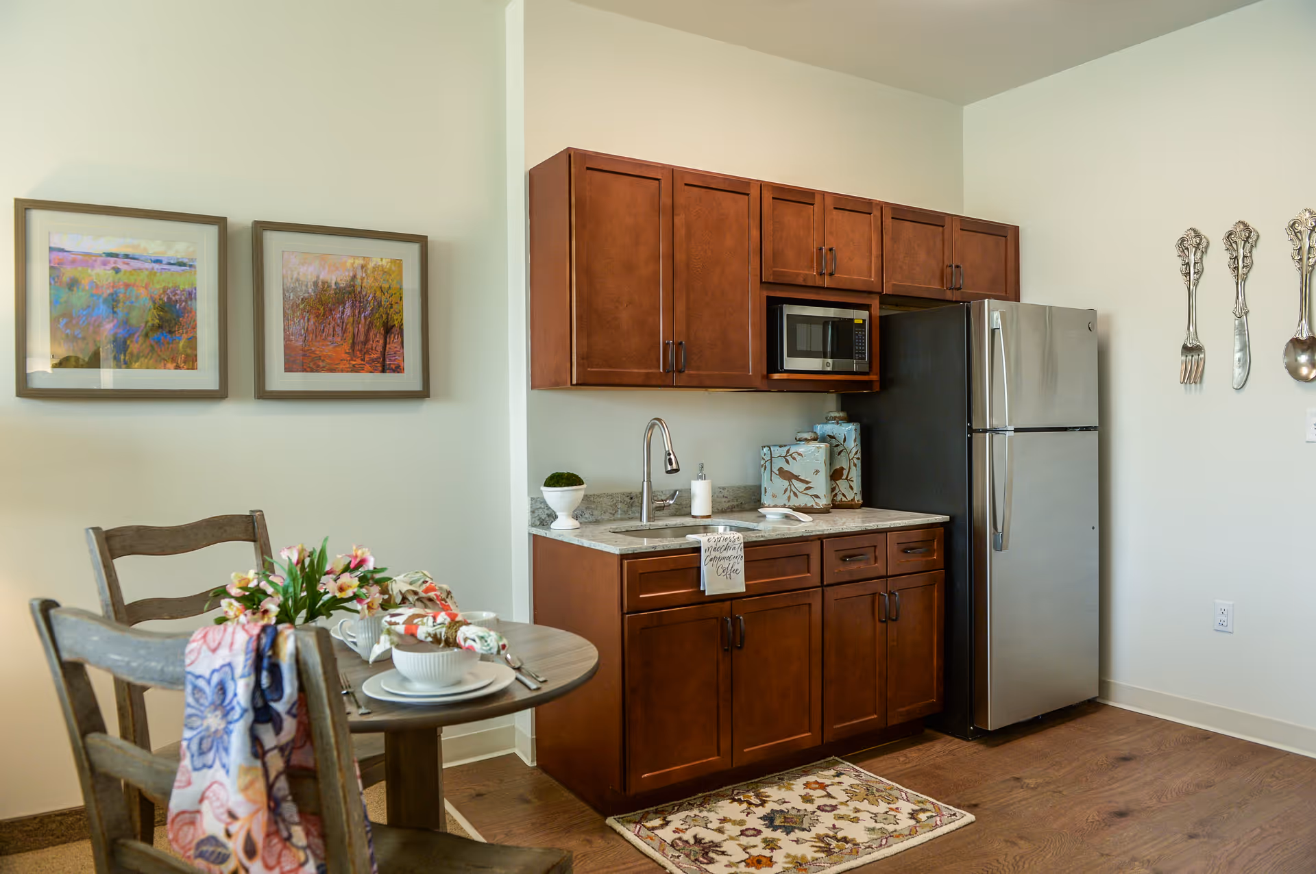 A small kitchen area with wooden cabinets, a stainless steel refrigerator, a microwave, and a sink. There is a small round dining table with two wooden chairs, set with white dishes, napkins, and a floral centerpiece. Two framed colorful landscape paintings hang on the wall, and decorative oversized silver utensils are mounted on another wall.