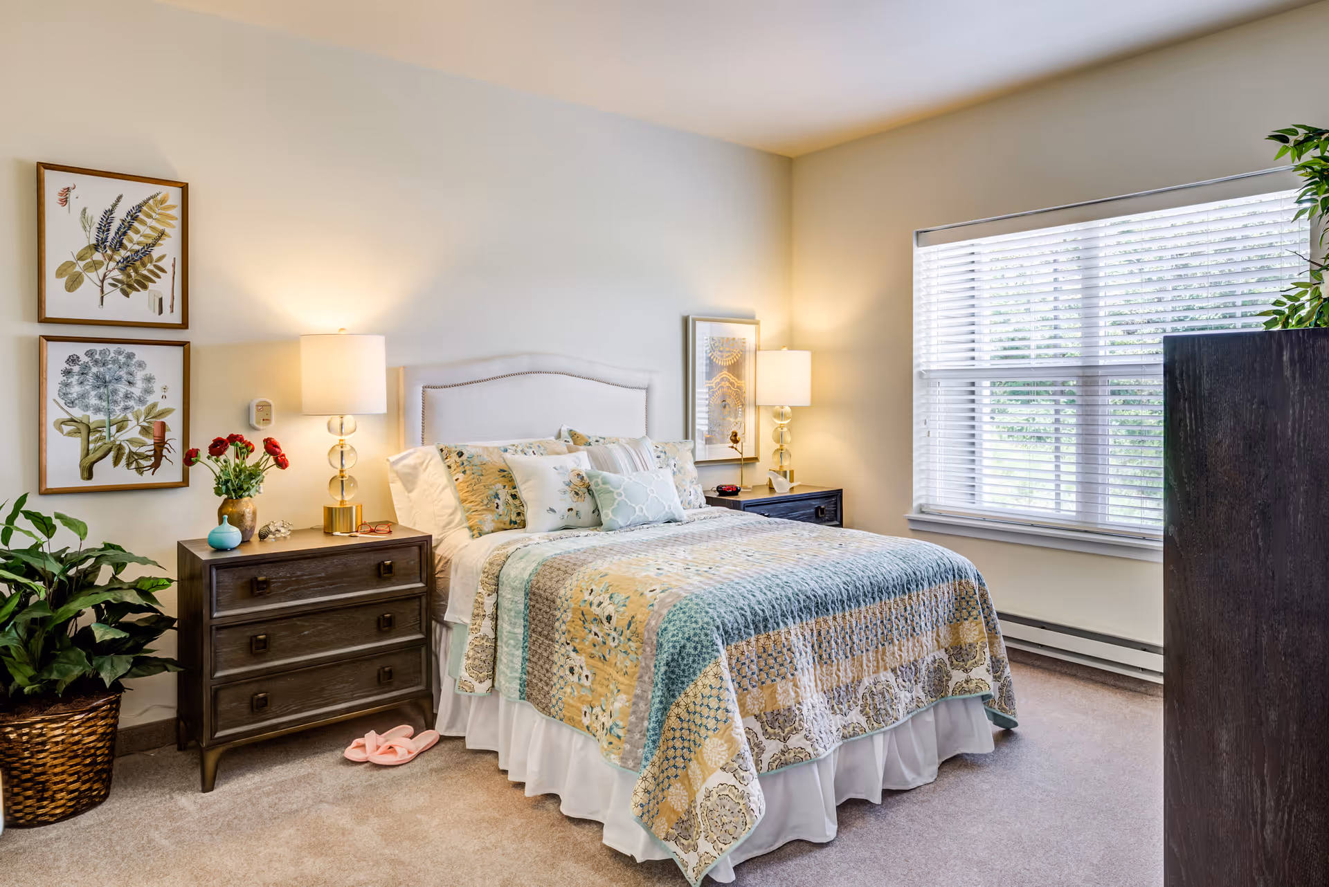 A cozy bedroom with a neatly made bed featuring a patterned quilt and multiple pillows. There are two bedside tables with lamps on each side of the bed. The room has light-colored walls adorned with framed botanical artwork. A large window with white blinds lets in natural light, and there is a potted plant in a woven basket on the floor next to one bedside table. Pink slippers are placed on the carpeted floor beside the bed.