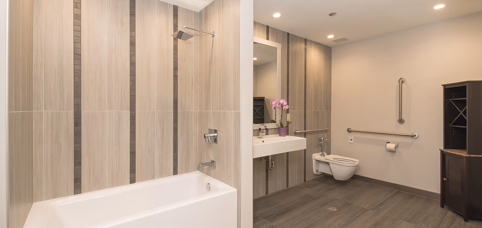 A modern bathroom with a white bathtub and overhead shower on the left, a wall-mounted sink with a mirror and a purple orchid on the counter in the center, and a wall-mounted toilet with grab bars on the right. The walls and floor have light brown and beige tiles, and there is a dark wooden cabinet in the corner.