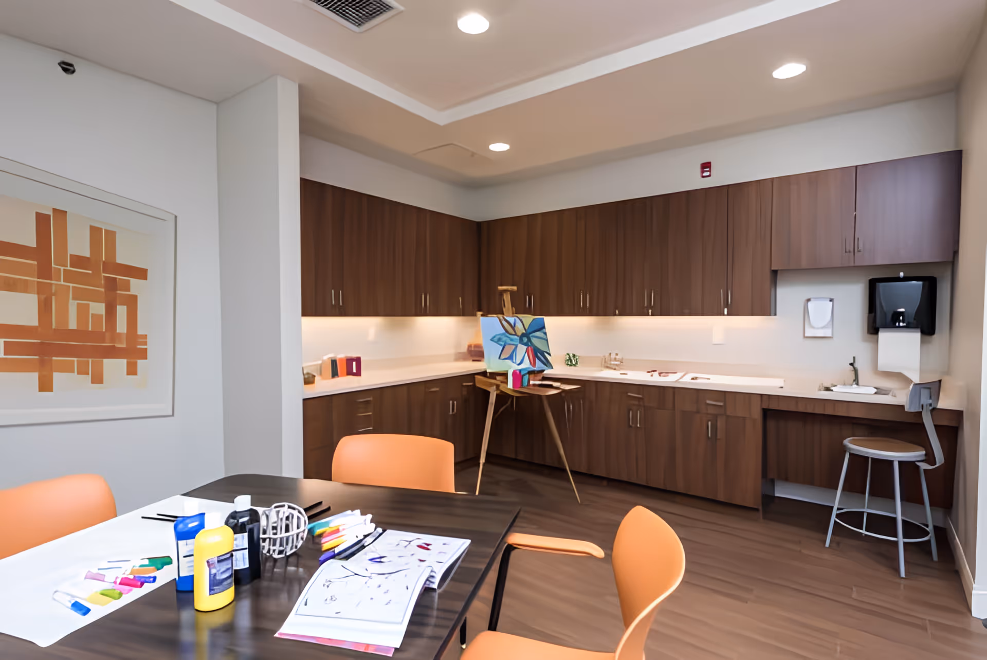 A well-lit art activity room with a table holding paint bottles, brushes, and coloring sheets. The room features orange chairs around the table, wooden cabinets along the walls, an easel with a painting of a flower, and a small sink area with a stool and paper towel dispenser.