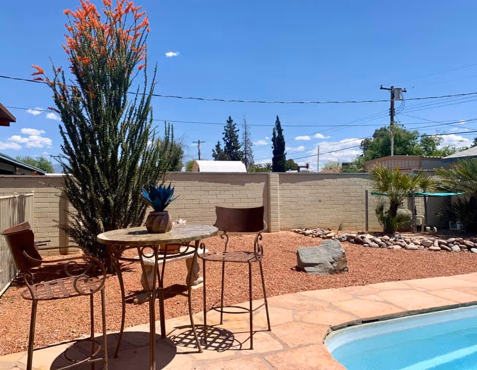 Outdoor patio area with a small round table and two metal chairs next to a swimming pool. The ground is covered with reddish gravel and there are various plants including a tall cactus and palm trees. A beige brick wall encloses the area under a clear blue sky.
