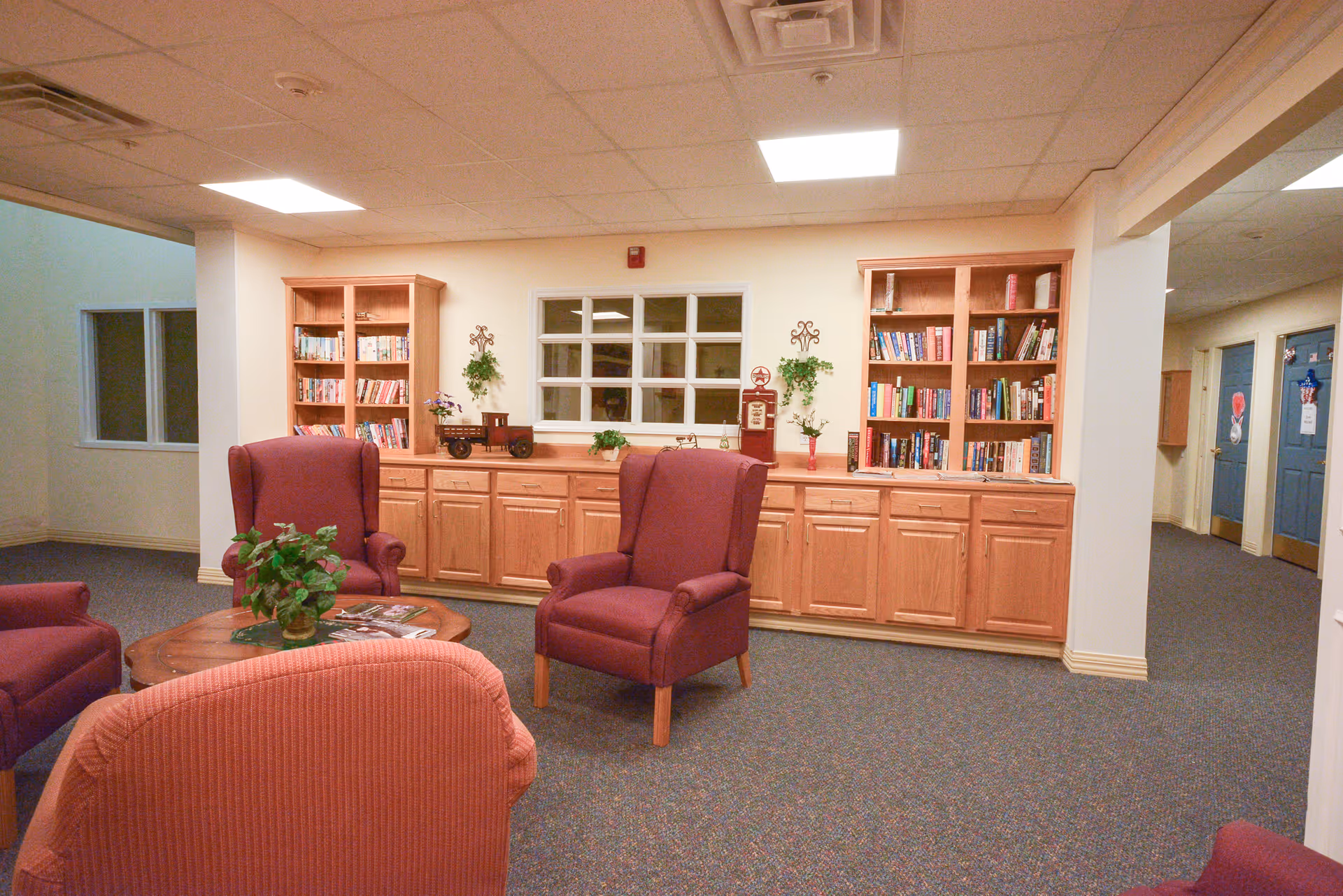 A cozy common area in an assisted living facility featuring several upholstered armchairs arranged around a wooden coffee table with a potted plant and magazines. Behind the seating area, there are built-in wooden cabinets and bookshelves filled with books and decorative items. The room has a carpeted floor and a drop ceiling with fluorescent lighting.
