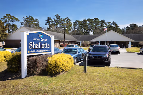 Exterior view of Autumn Care of Shallotte Health & Rehabilitation Center with a large sign in the foreground, several parked cars, a flagpole with an American flag, and a building surrounded by trees under a clear blue sky.