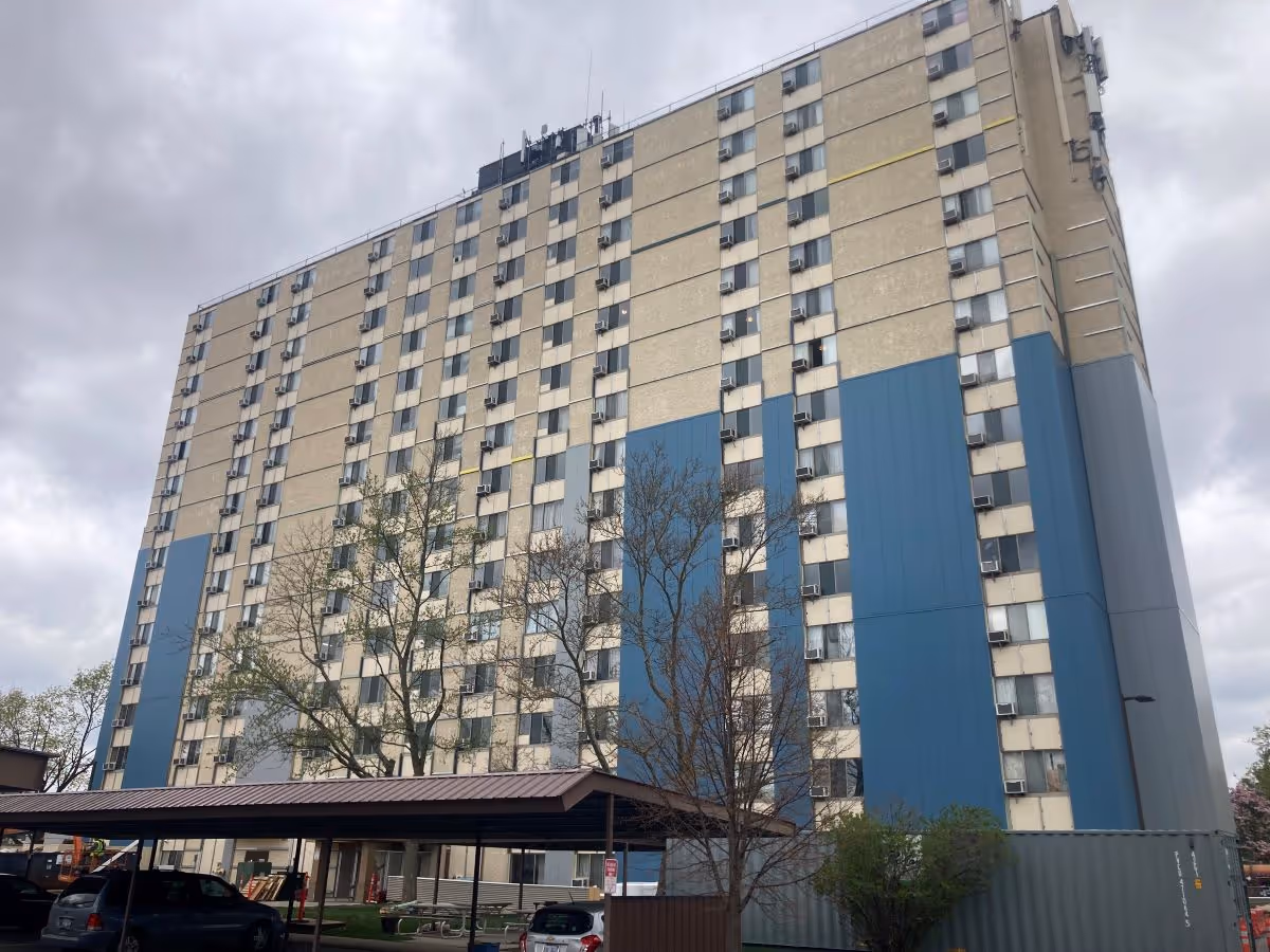 Tall multi-story residential building with blue vertical panels, many windows with window air conditioners, and a covered parking area in front under a cloudy sky.