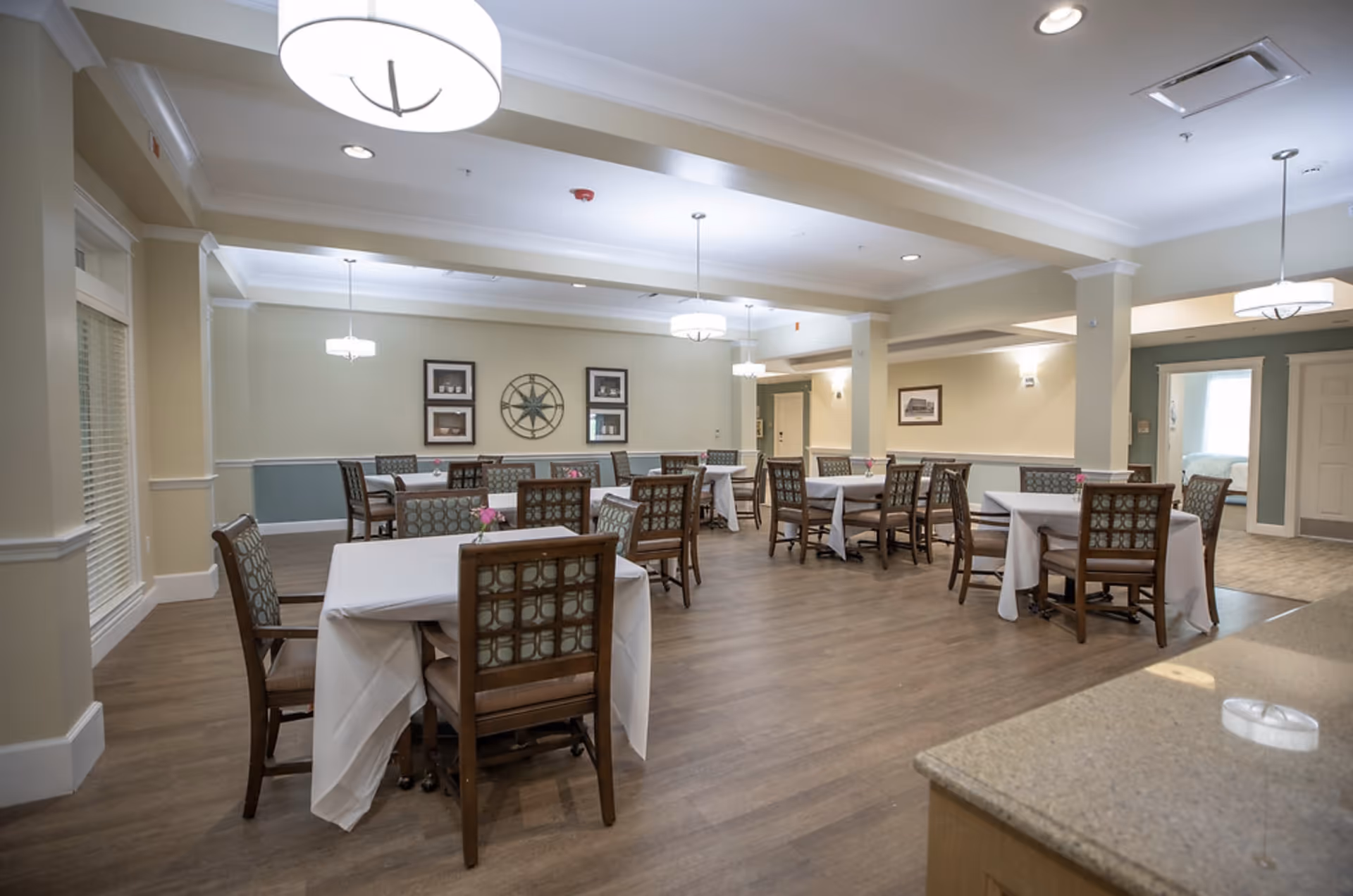 A spacious dining room with several tables covered with white tablecloths and surrounded by wooden chairs with patterned cushions. The room has light-colored walls with decorative molding, framed artwork, and modern pendant lights hanging from the ceiling. The floor is wood, and there is a countertop visible in the foreground.